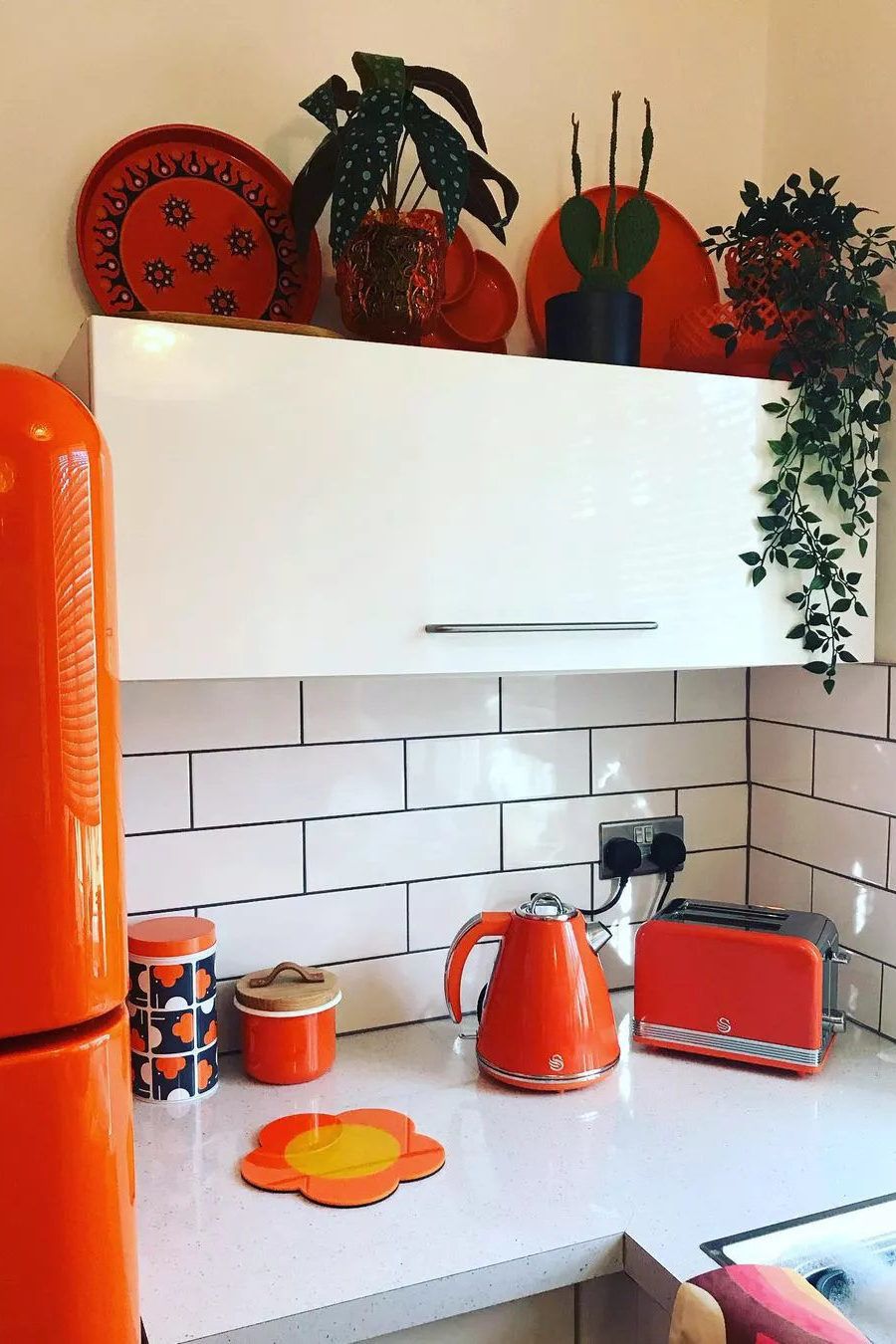 Bright orange glossy kettle, toaster, and fridge in glossy white kitchen with subway style white backsplash and orange decorative plates on top of glossy white cabinets. 