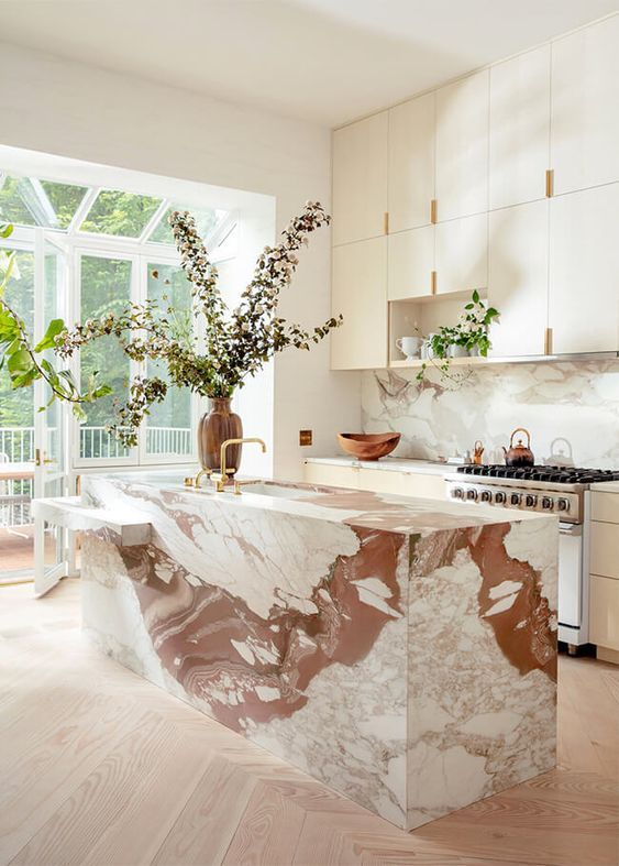 light hardwood floor, terracotta dn white marble island in white kitchen with large floor to ceiling windows and large vase with green branches.