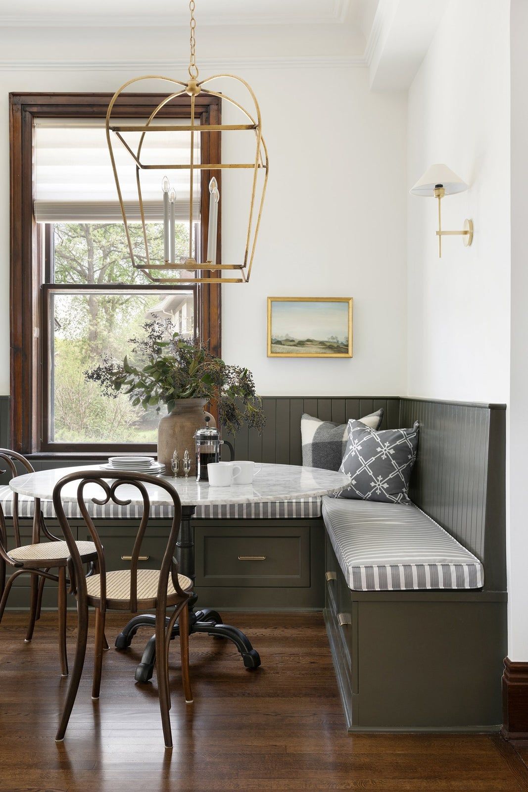 breakfast nook with large gold and glass candle style pendant over marble top table, chaki bench, dark hardwood floor and french bistro chairs and window frame.